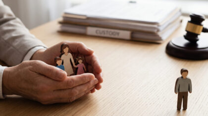 Primer plano de manos profesionales sosteniendo simbólicas figuras de madera de una familia en un escritorio legal. Al fondo, un mazo de juez y archivos etiquetados representan una decisión de custodia exclusiva.